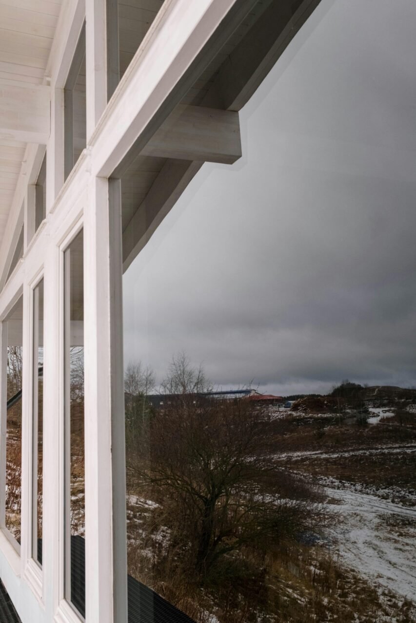 Fotografia de detalhe de fachada envidraçada branca refletindo a paisagem exterior de inverno, com árvores despidas e colinas parcialmente cobertas de neve em Lisboa.