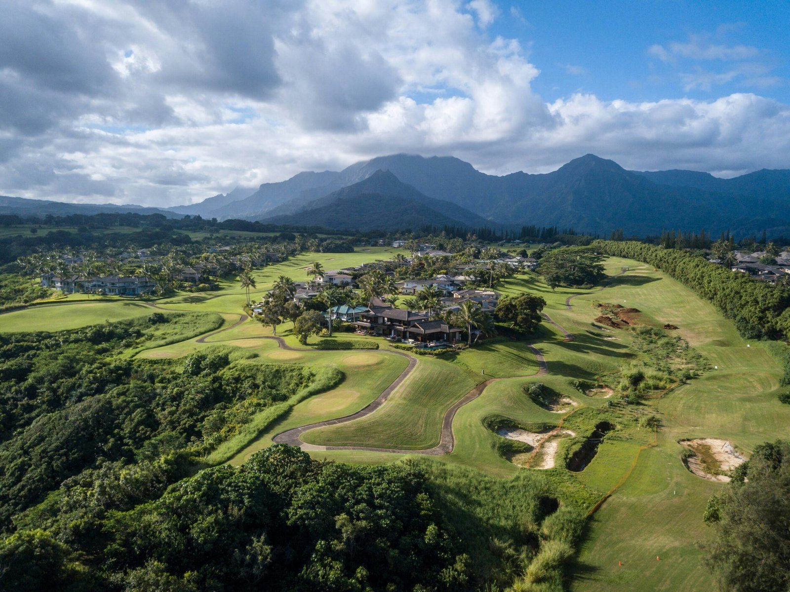 Fotografia aérea de um campo de golfe com fairways ondulados, casas ao centro e uma cordilheira de montanhas ao fundo sob céu parcialmente nublado.