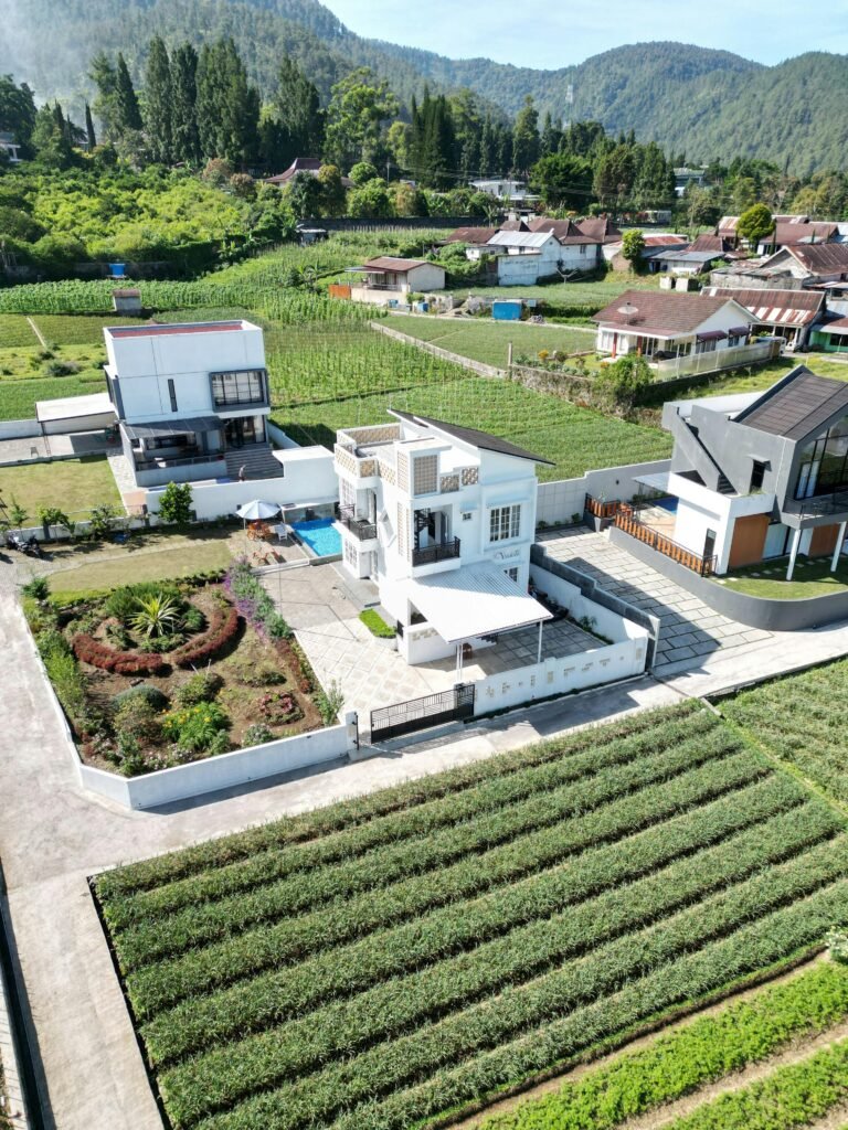 Fotografia aérea de um imóvel moderno de dois andares com jardim paisagístico e piscina, rodeado por campos agrícolas verdes e colinas arborizadas ao fundo.